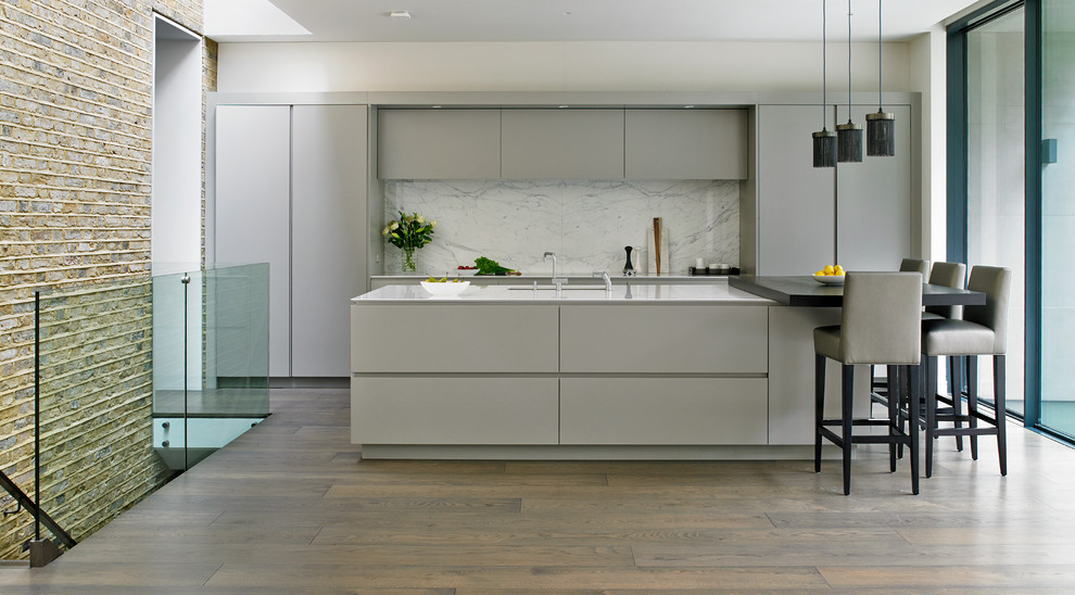Contemporary kitchen in London with flat-panel cabinets, grey cabinets, composite countertops, white splashback, stone slab splashback, black appliances, medium hardwood flooring and an island.