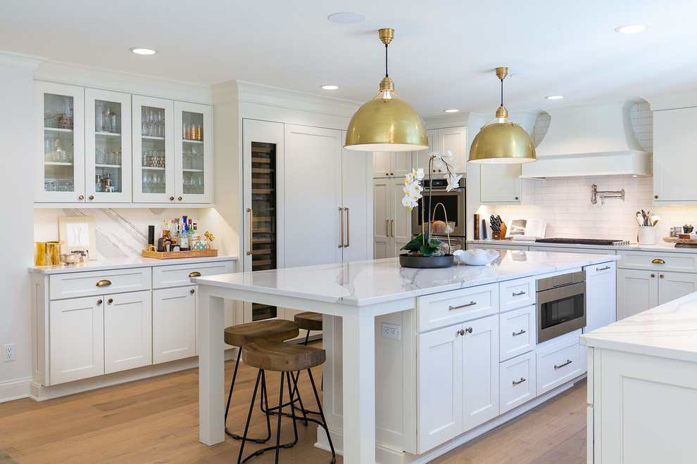 Photo of a large traditional u-shaped kitchen/diner in Kansas City with shaker cabinets, white cabinets, engineered stone countertops, white splashback, metro tiled splashback, integrated appliances, an island, white worktops, medium hardwood flooring and brown floors.