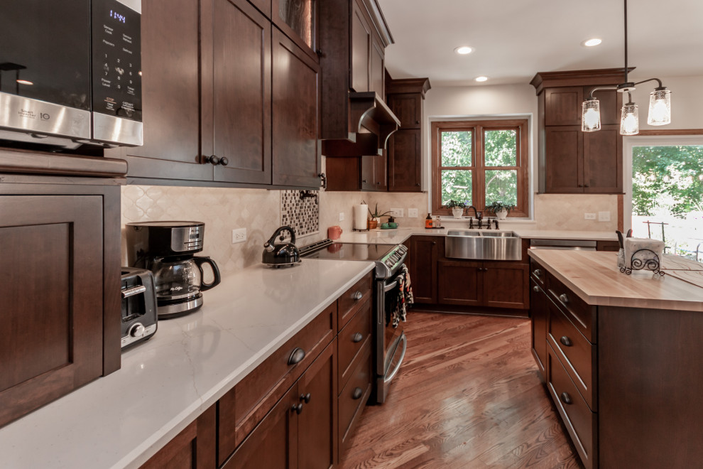 Large classic l-shaped kitchen/diner in Chicago with a belfast sink, shaker cabinets, brown cabinets, engineered stone countertops, beige splashback, marble splashback, stainless steel appliances, medium hardwood flooring, an island, brown floors and beige worktops.