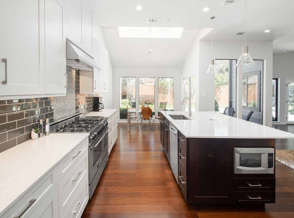 Photo of a large classic single-wall open plan kitchen in Dallas with a submerged sink, flat-panel cabinets, white cabinets, engineered stone countertops, grey splashback, glass tiled splashback, stainless steel appliances, bamboo flooring, an island, brown floors and white worktops.