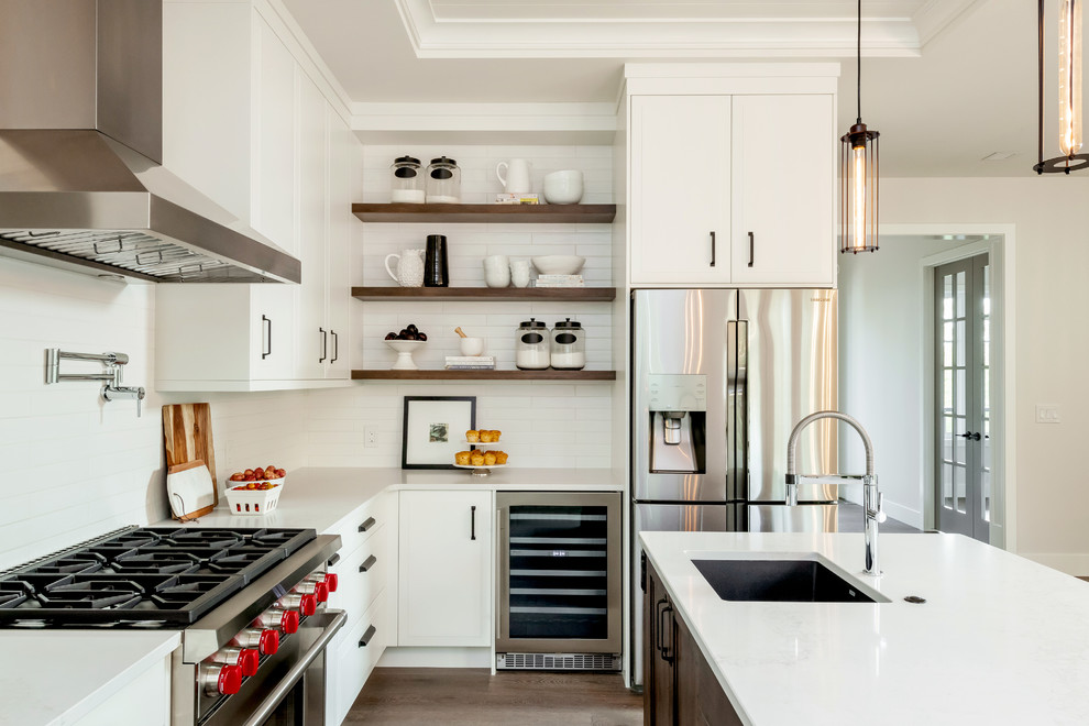 Design ideas for a large traditional l-shaped kitchen in Other with a belfast sink, raised-panel cabinets, white cabinets, engineered stone countertops, white splashback, metro tiled splashback, stainless steel appliances, medium hardwood flooring, an island, brown floors and white worktops.