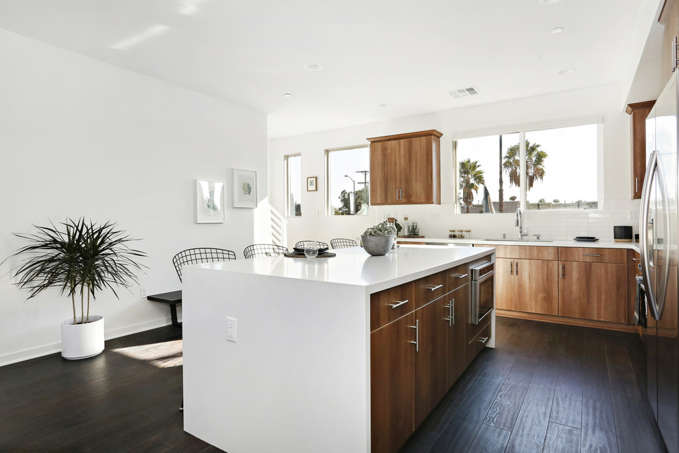 Inspiration for a contemporary l-shaped open plan kitchen in Los Angeles with a submerged sink, flat-panel cabinets, medium wood cabinets, white splashback, metro tiled splashback, stainless steel appliances, dark hardwood flooring, brown floors and white worktops.