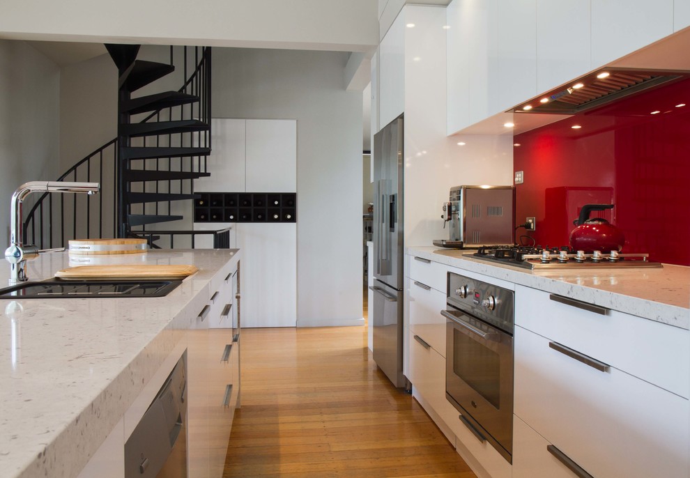 Modern inredning av ett mellanstort kök, med släta luckor, vita skåp, bänkskiva i kvarts, rött stänkskydd, glaspanel som stänkskydd, rostfria vitvaror, mellanmörkt trägolv och en köksö