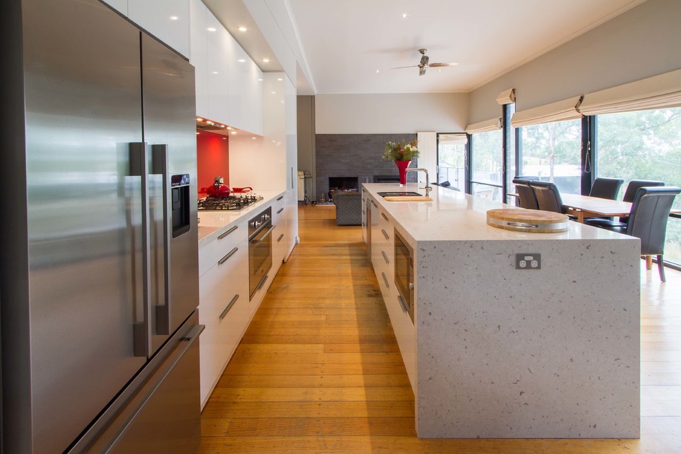This is an example of a medium sized contemporary galley open plan kitchen in Melbourne with flat-panel cabinets, white cabinets, engineered stone countertops, red splashback, glass sheet splashback, stainless steel appliances, medium hardwood flooring and an island.