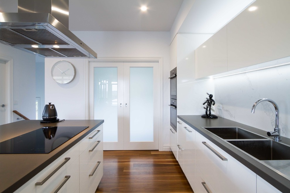 Large modern galley open plan kitchen in Melbourne with a built-in sink, flat-panel cabinets, white cabinets, engineered stone countertops, stone slab splashback, stainless steel appliances, medium hardwood flooring and an island.