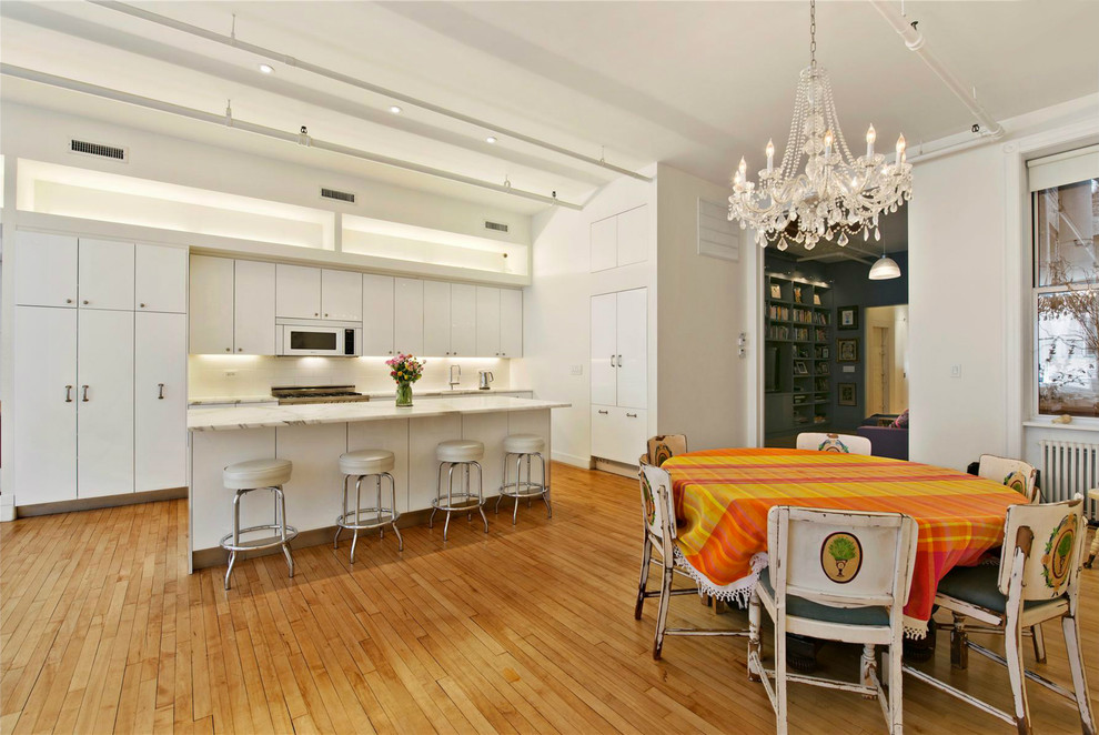This is an example of a medium sized industrial kitchen in New York with flat-panel cabinets, white cabinets, white splashback, ceramic splashback, light hardwood flooring, an island and a submerged sink.