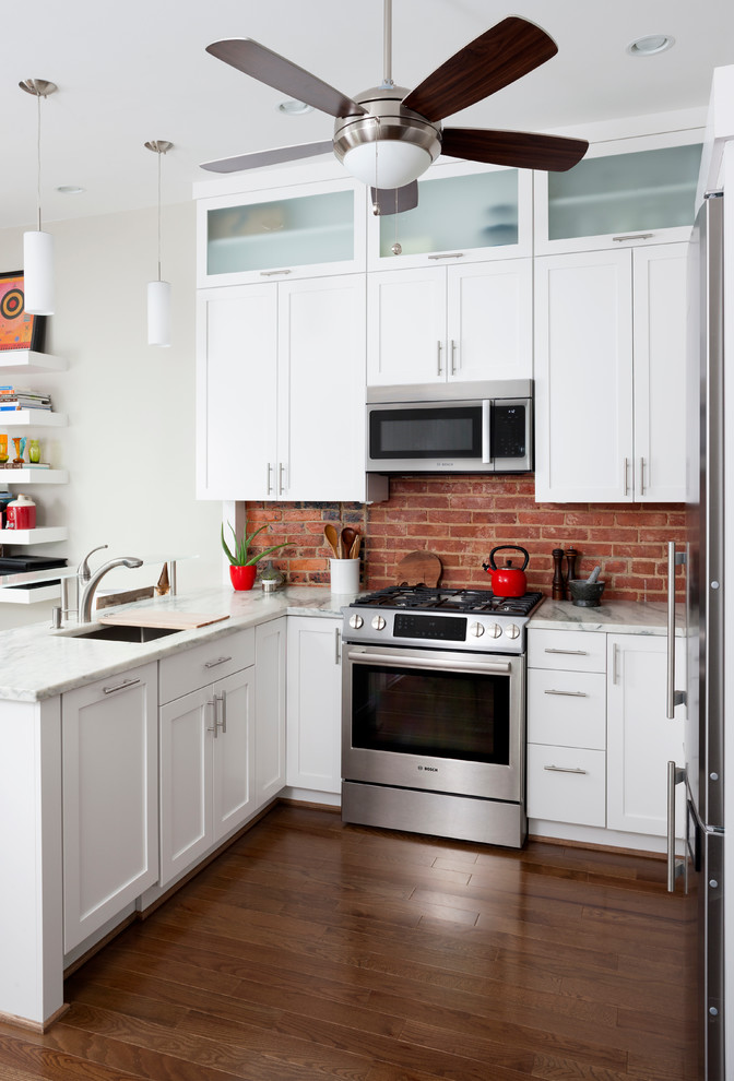 Klassisk inredning av ett litet kök, med vita skåp, rött stänkskydd, rostfria vitvaror och mellanmörkt trägolv