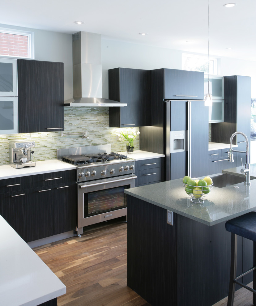 Photo of a medium sized contemporary l-shaped kitchen/diner in Denver with a belfast sink, flat-panel cabinets, black cabinets, granite worktops, green splashback, glass tiled splashback, stainless steel appliances, medium hardwood flooring, an island and brown floors.