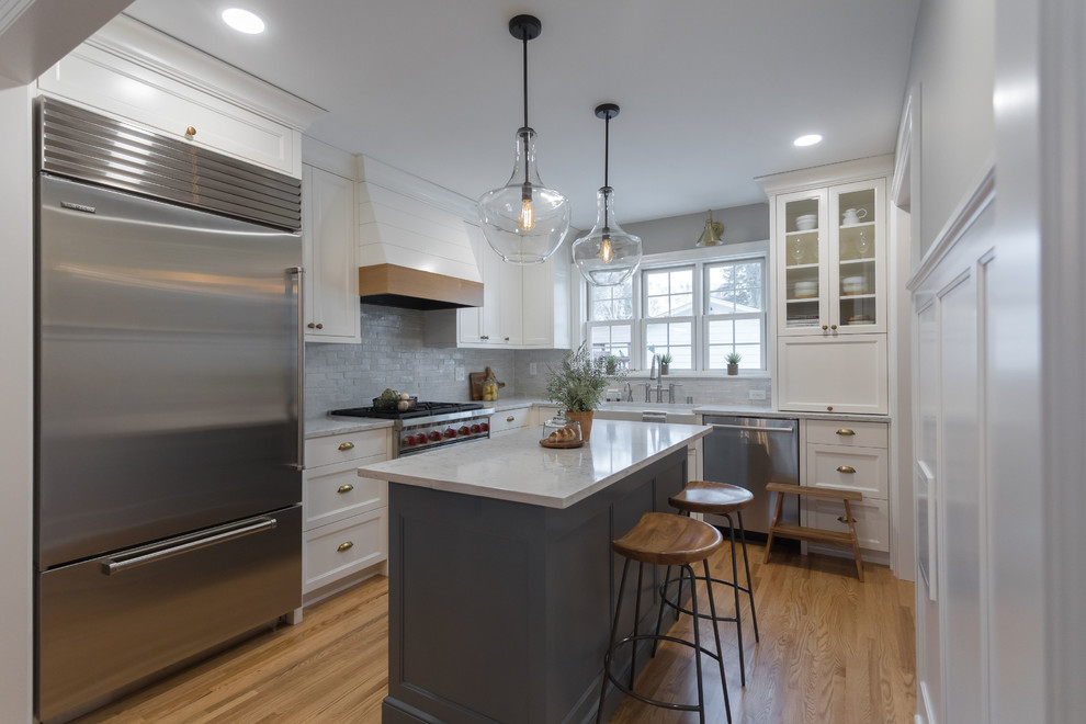 Photo of a traditional l-shaped kitchen in Minneapolis with a belfast sink, recessed-panel cabinets, white cabinets, stainless steel appliances, medium hardwood flooring, an island, brown floors and white worktops.