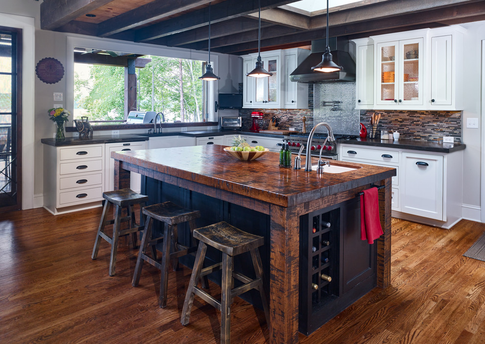 This is an example of a rustic l-shaped kitchen in Other with a belfast sink, white cabinets, multi-coloured splashback, stainless steel appliances, medium hardwood flooring, an island and black worktops.