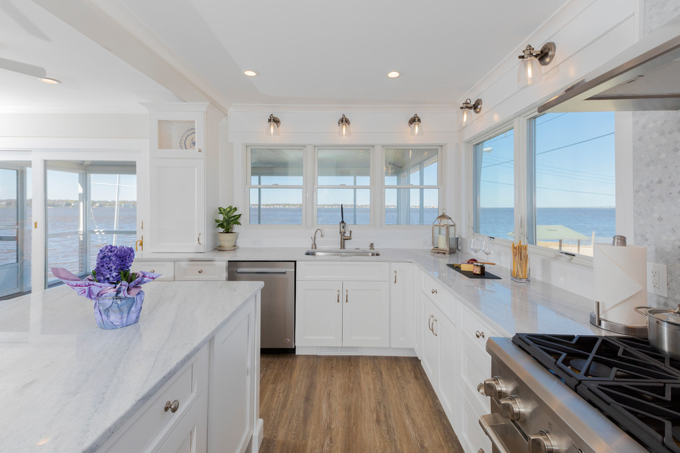 Photo of a medium sized nautical u-shaped kitchen/diner in New York with a submerged sink, shaker cabinets, white cabinets, quartz worktops, grey splashback, marble splashback, stainless steel appliances, painted wood flooring, an island, brown floors and white worktops.