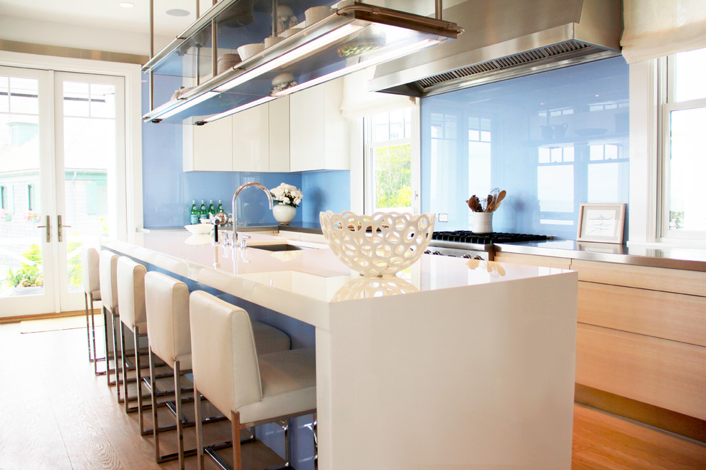 Foto på ett maritimt kök, med släta luckor, blått stänkskydd, glaspanel som stänkskydd, rostfria vitvaror, ljust trägolv, en undermonterad diskho, bänkskiva i rostfritt stål och en köksö
