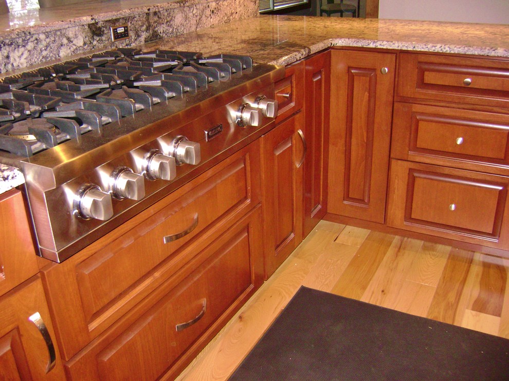 Klassisk inredning av ett kök, med luckor med upphöjd panel, skåp i mörkt trä, granitbänkskiva, grått stänkskydd, rostfria vitvaror, ljust trägolv och en halv köksö