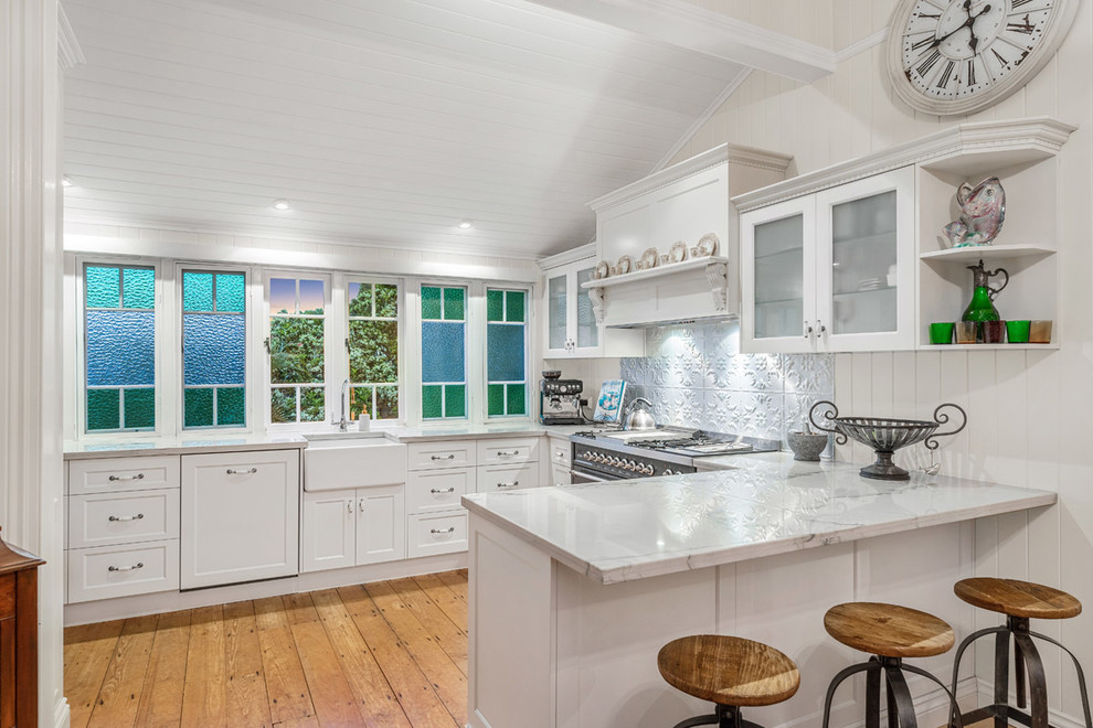 Klassisk inredning av ett mellanstort vit vitt kök, med en rustik diskho, luckor med glaspanel, vita skåp, bänkskiva i kvartsit, stänkskydd med metallisk yta och en köksö
