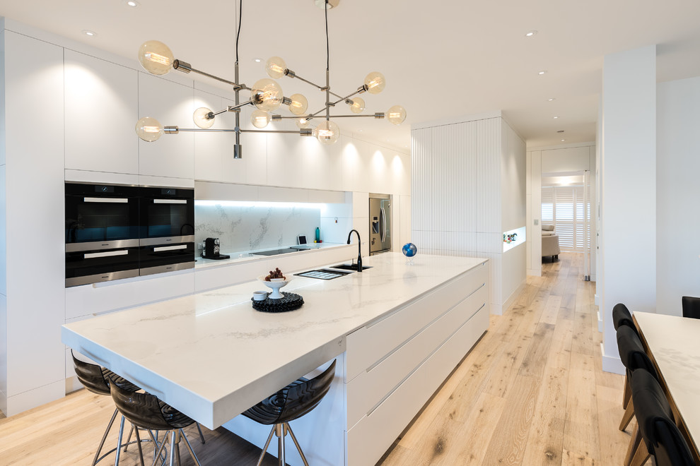 Photo of a contemporary kitchen/diner in Adelaide with a submerged sink, flat-panel cabinets, white cabinets, white splashback, stainless steel appliances, light hardwood flooring, an island, beige floors and white worktops.