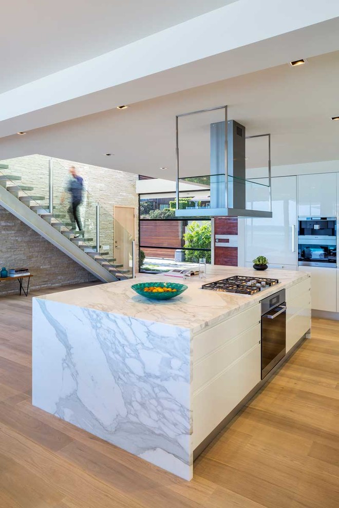 Medium sized contemporary galley open plan kitchen in Los Angeles with flat-panel cabinets, white cabinets, marble worktops, white splashback, an island, light hardwood flooring and stainless steel appliances.