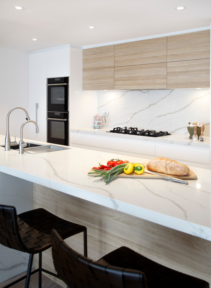 Inspiration for a large modern galley open plan kitchen in Sydney with a submerged sink, flat-panel cabinets, white cabinets, engineered stone countertops, white splashback, stone slab splashback, black appliances, light hardwood flooring, an island and white worktops.