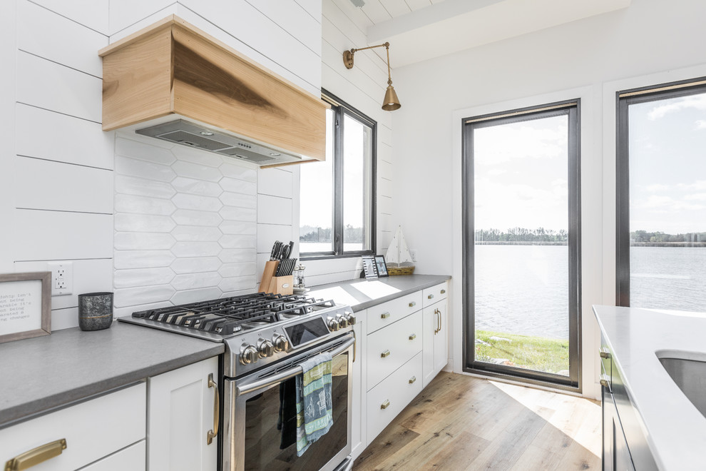 Foto di un cucina con isola centrale costiero con ante in stile shaker, ante bianche, elettrodomestici in acciaio inossidabile, parquet chiaro e top grigio