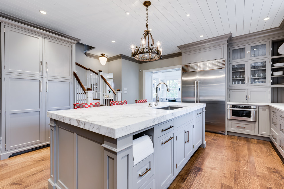 Klassisk inredning av ett kök, med en undermonterad diskho, luckor med infälld panel, grå skåp, marmorbänkskiva, vitt stänkskydd, rostfria vitvaror, mellanmörkt trägolv och en köksö