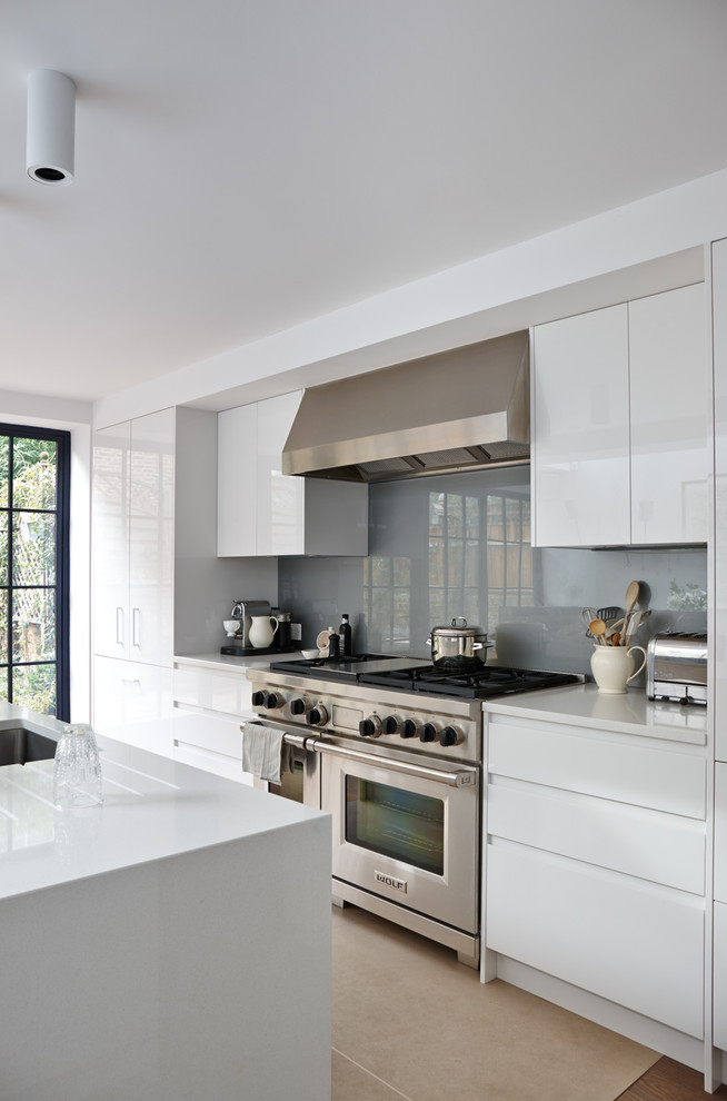 Medium sized modern grey and cream galley open plan kitchen in London with an integrated sink, flat-panel cabinets, white cabinets, composite countertops, grey splashback, glass sheet splashback, stainless steel appliances, medium hardwood flooring, an island and white worktops.