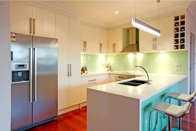 Modern inredning av ett kök, med en undermonterad diskho, luckor med infälld panel, vita skåp, grönt stänkskydd, glaspanel som stänkskydd och rostfria vitvaror