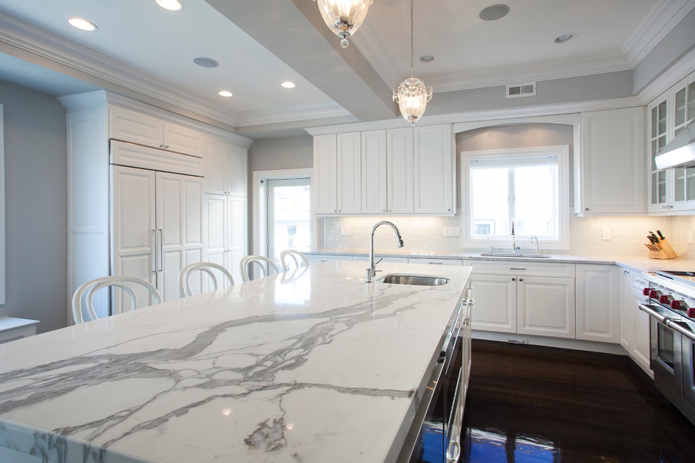 Example of a mid-sized classic l-shaped dark wood floor eat-in kitchen design in New York with an undermount sink, raised-panel cabinets, white cabinets, quartz countertops, white backsplash, mosaic tile backsplash, stainless steel appliances, an island and white countertops