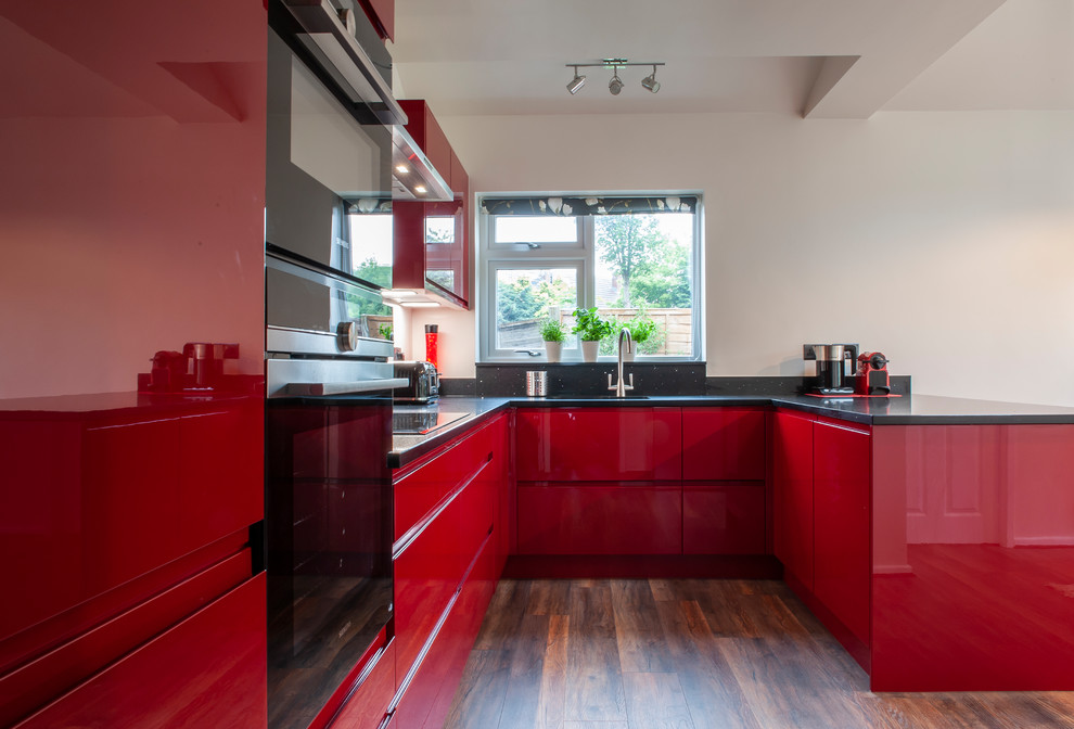Große Moderne Küche in U-Form mit roten Schränken, Quarzit-Arbeitsplatte, Laminat, braunem Boden, schwarzer Arbeitsplatte, flächenbündigen Schrankfronten, schwarzen Elektrogeräten und Halbinsel in Cheshire