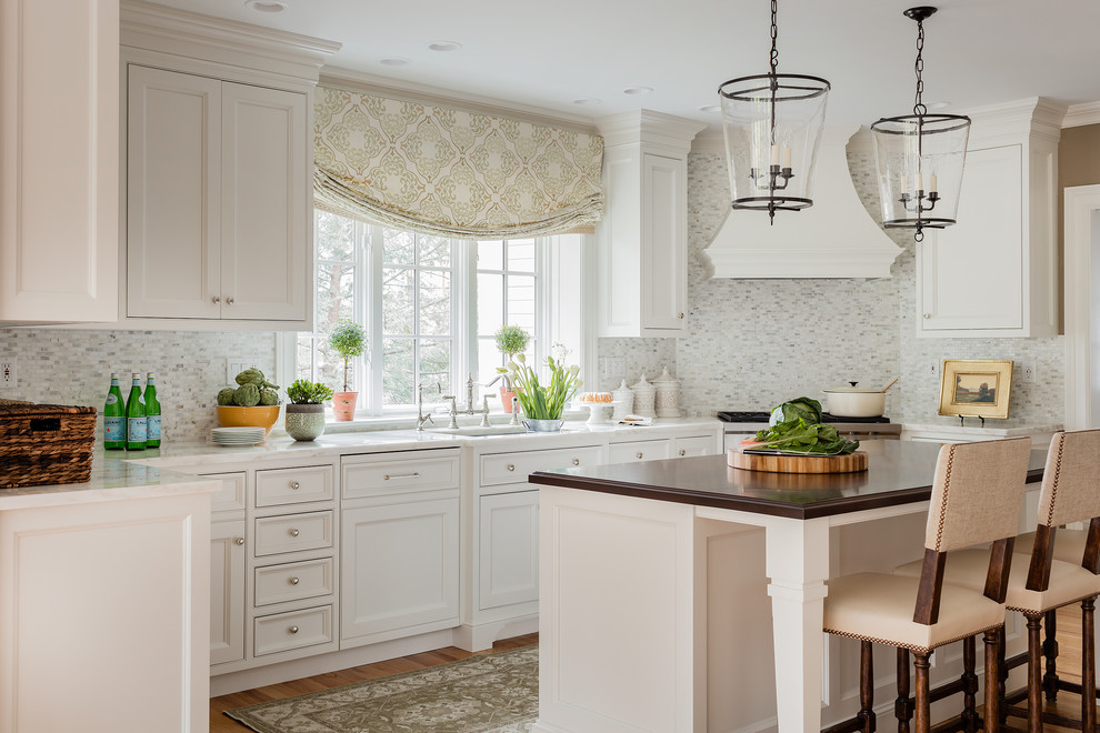 Inredning av ett klassiskt mellanstort kök, med en undermonterad diskho, luckor med profilerade fronter, vita skåp, marmorbänkskiva, vitt stänkskydd, stänkskydd i mosaik, integrerade vitvaror, ljust trägolv och en köksö