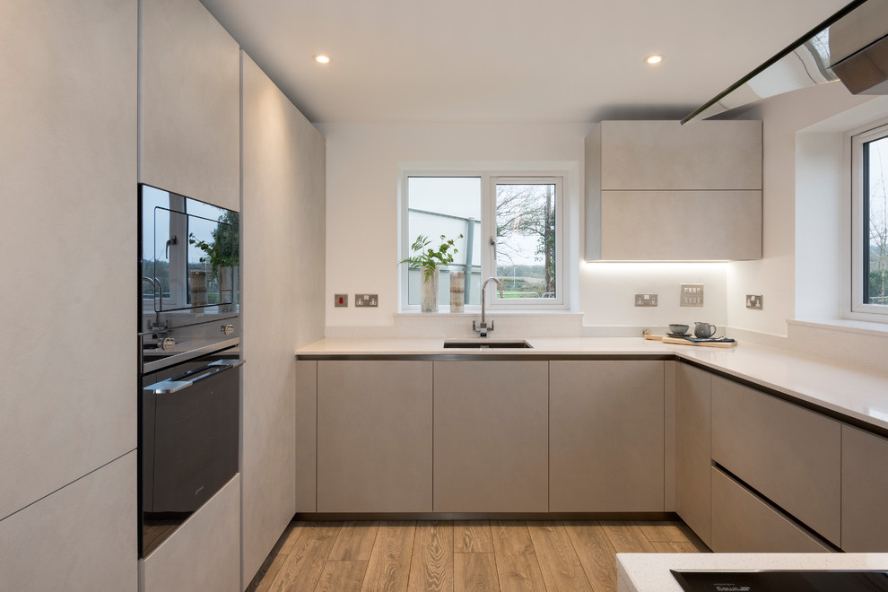 Mittelgroße Moderne Wohnküche mit Waschbecken, Glasfronten, blauen Schränken, Küchenrückwand in Rot, Küchengeräten aus Edelstahl, Laminat, grauem Boden und weißer Arbeitsplatte in Devon