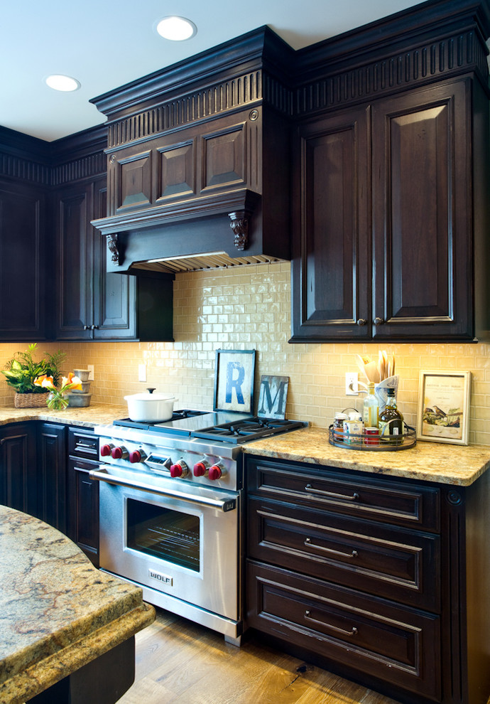 Example of a mid-sized classic l-shaped medium tone wood floor enclosed kitchen design in St Louis with a farmhouse sink, raised-panel cabinets, black cabinets, granite countertops, beige backsplash, subway tile backsplash and stainless steel appliances