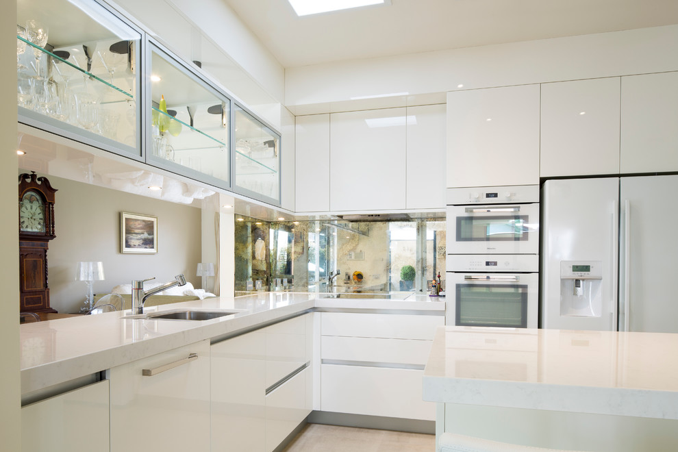 Bild på ett mellanstort funkis kök, med en undermonterad diskho, vita skåp, bänkskiva i kvarts, spegel som stänkskydd, vita vitvaror och en köksö