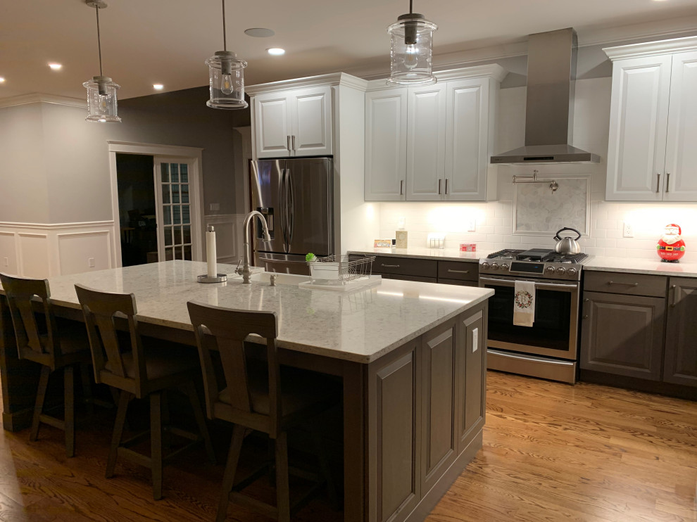 Design ideas for a medium sized classic l-shaped kitchen pantry in Providence with a belfast sink, raised-panel cabinets, white cabinets, engineered stone countertops, white splashback, porcelain splashback, stainless steel appliances, medium hardwood flooring, an island, brown floors and white worktops.