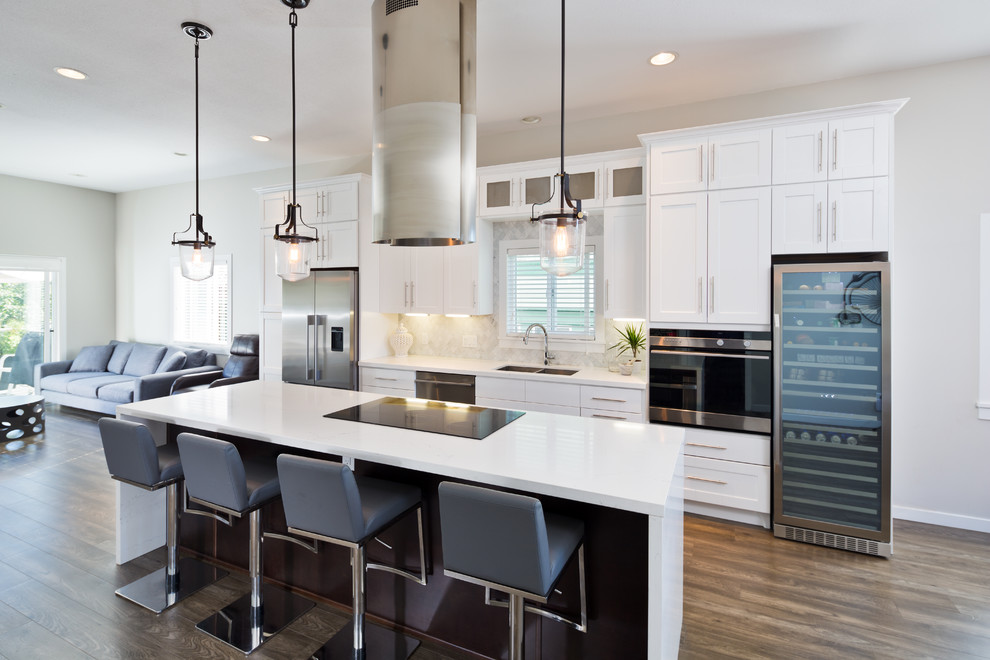 Transitional galley medium tone wood floor kitchen photo in Vancouver with a double-bowl sink, shaker cabinets, white cabinets, white backsplash, stainless steel appliances and an island
