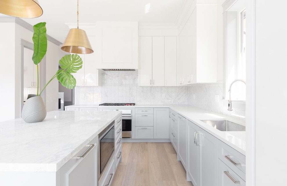Photo of a medium sized traditional l-shaped open plan kitchen in Vancouver with a single-bowl sink, shaker cabinets, white cabinets, marble worktops, white splashback, marble splashback, integrated appliances, light hardwood flooring and an island.