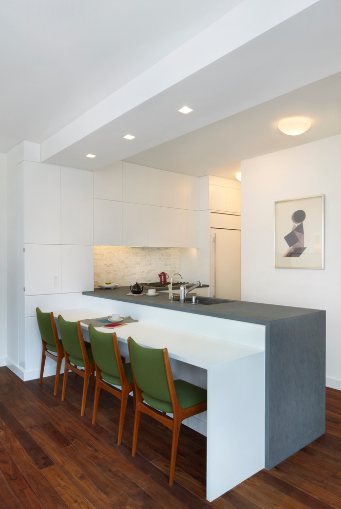 Inspiration for a small modern l-shaped medium tone wood floor eat-in kitchen remodel in New York with an undermount sink, flat-panel cabinets, white cabinets, limestone countertops, stone tile backsplash, paneled appliances, a peninsula and white backsplash