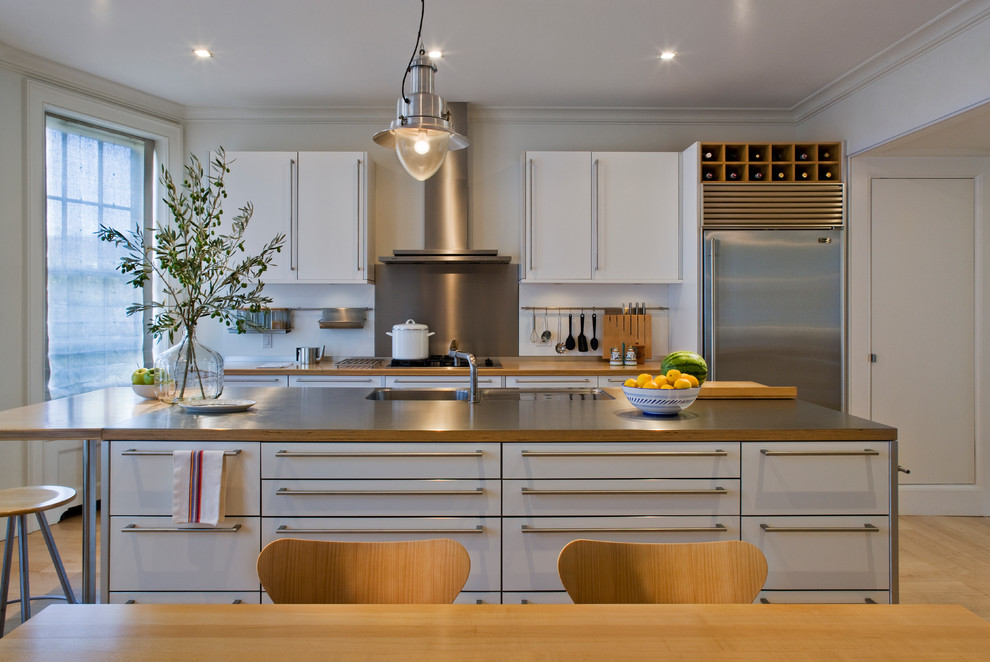 Foto på ett mellanstort nordiskt grå linjärt kök och matrum, med en undermonterad diskho, släta luckor, vita skåp, bänkskiva i rostfritt stål, stänkskydd med metallisk yta, rostfria vitvaror, ljust trägolv, en köksö och brunt golv
