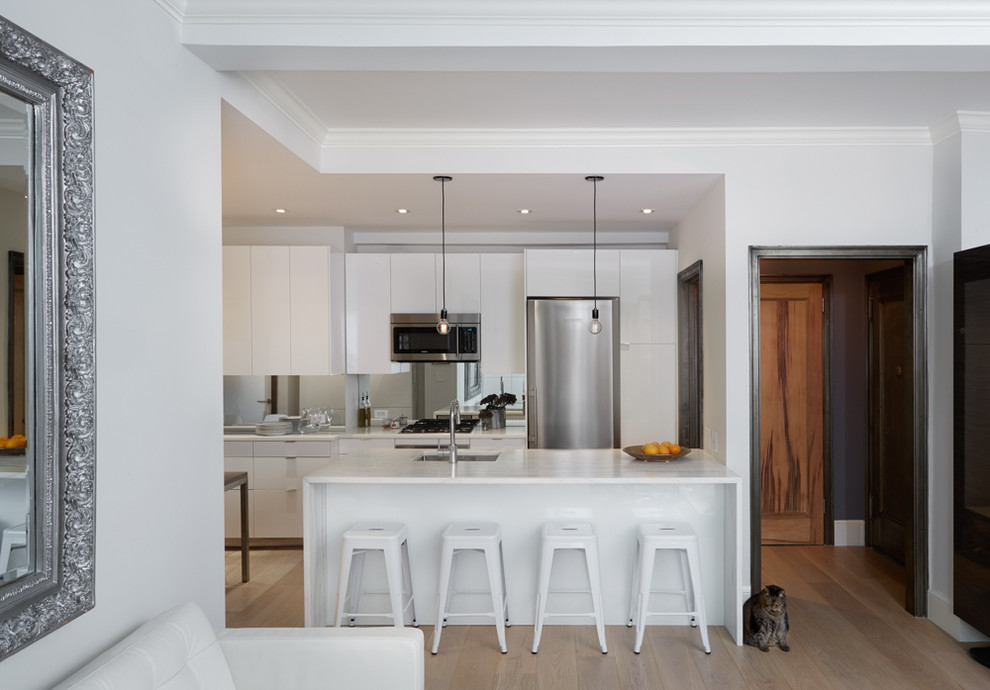 Inredning av ett modernt litet kök, med en undermonterad diskho, släta luckor, vita skåp, marmorbänkskiva, stänkskydd med metallisk yta, spegel som stänkskydd, rostfria vitvaror, ljust trägolv, en halv köksö och beiget golv