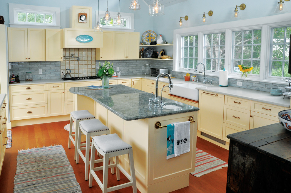 Example of a beach style medium tone wood floor and brown floor kitchen design in Boston with a farmhouse sink, shaker cabinets, yellow cabinets, gray backsplash, an island, gray countertops, granite countertops and ceramic backsplash