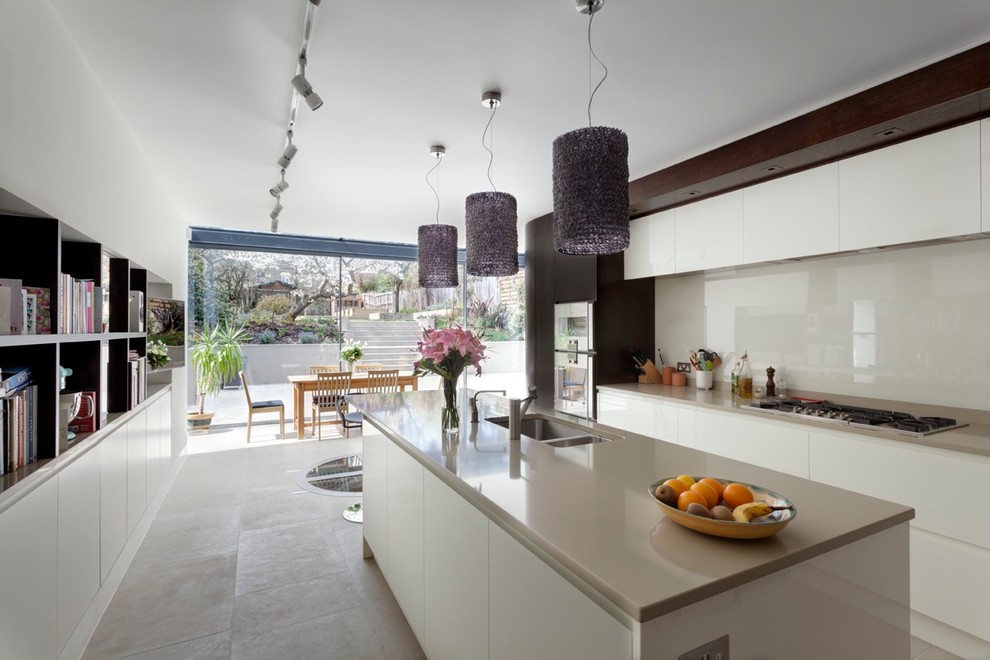 Photo of a large contemporary l-shaped kitchen/diner in London with a built-in sink, flat-panel cabinets, white cabinets, composite countertops, white splashback, glass sheet splashback, stainless steel appliances, limestone flooring, an island, grey floors and grey worktops.