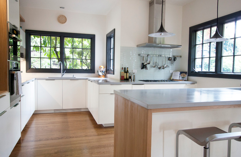 Example of a large minimalist u-shaped light wood floor eat-in kitchen design in Portland with an undermount sink, flat-panel cabinets, white cabinets, concrete countertops, white backsplash, glass tile backsplash, stainless steel appliances and a peninsula