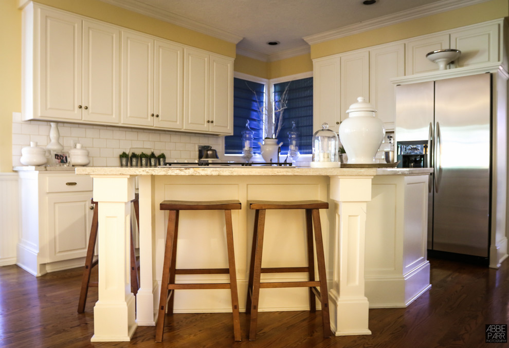 Klassisk inredning av ett mellanstort kök, med mörkt trägolv, en dubbel diskho, vitt stänkskydd, stänkskydd i tunnelbanekakel, rostfria vitvaror, luckor med upphöjd panel, vita skåp, granitbänkskiva och en köksö