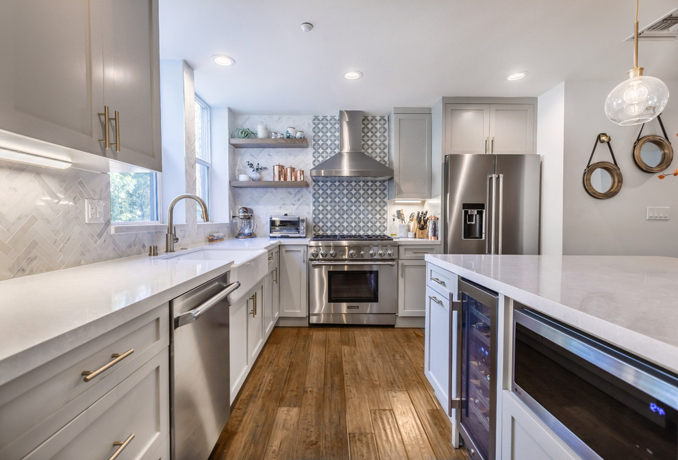 Inspiration for a medium sized country l-shaped kitchen/diner in Los Angeles with a belfast sink, shaker cabinets, grey cabinets, engineered stone countertops, white splashback, marble splashback, stainless steel appliances, medium hardwood flooring, an island and brown floors.