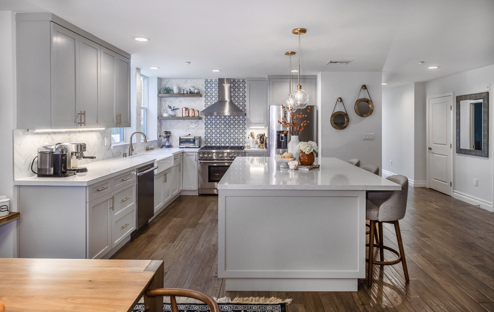 Design ideas for a medium sized country l-shaped kitchen/diner in Los Angeles with a belfast sink, shaker cabinets, grey cabinets, engineered stone countertops, white splashback, marble splashback, stainless steel appliances, medium hardwood flooring, an island and brown floors.