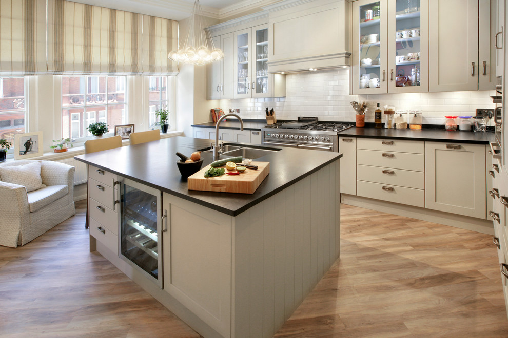 This is an example of a classic l-shaped kitchen in London with a double-bowl sink, glass-front cabinets, grey cabinets, white splashback, metro tiled splashback, stainless steel appliances, light hardwood flooring and an island.