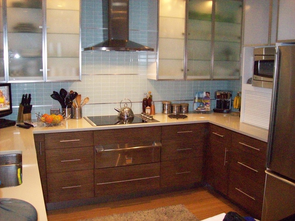 Inredning av ett 50 tals litet kök, med en undermonterad diskho, luckor med glaspanel, skåp i mellenmörkt trä, bänkskiva i kvarts, stänkskydd i glaskakel, rostfria vitvaror, bambugolv, blått stänkskydd, en halv köksö och brunt golv