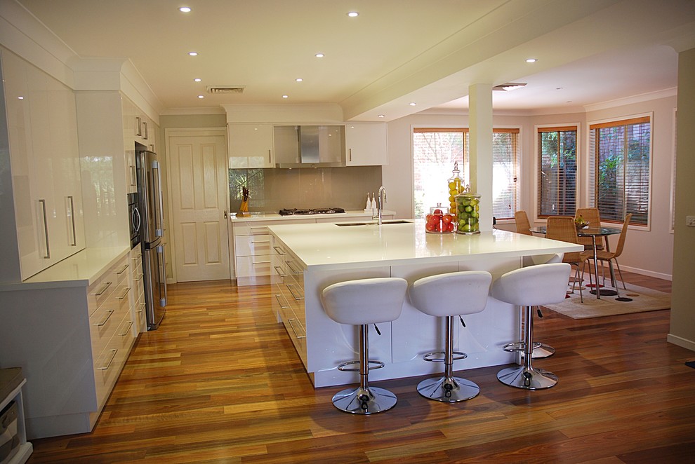Large modern l-shaped kitchen/diner in Sydney with a submerged sink, flat-panel cabinets, white cabinets, composite countertops, green splashback, glass sheet splashback, stainless steel appliances, medium hardwood flooring and an island.