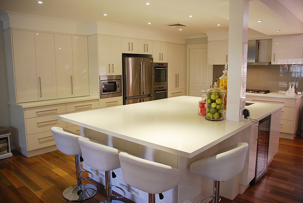 Large modern l-shaped kitchen/diner in Sydney with a submerged sink, flat-panel cabinets, white cabinets, composite countertops, brown splashback, glass sheet splashback, stainless steel appliances, medium hardwood flooring and an island.