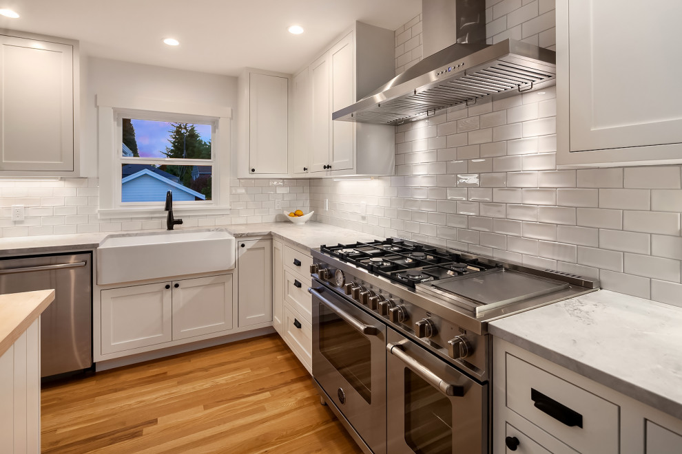 シアトルにある中くらいなトラディショナルスタイルのおしゃれなアイランドキッチン (エプロンフロントシンク、シェーカースタイル扉のキャビネット、白いキャビネット、珪岩カウンター、白いキッチンパネル、サブウェイタイルのキッチンパネル、シルバーの調理設備、淡色無垢フローリング、グレーのキッチンカウンター) の写真