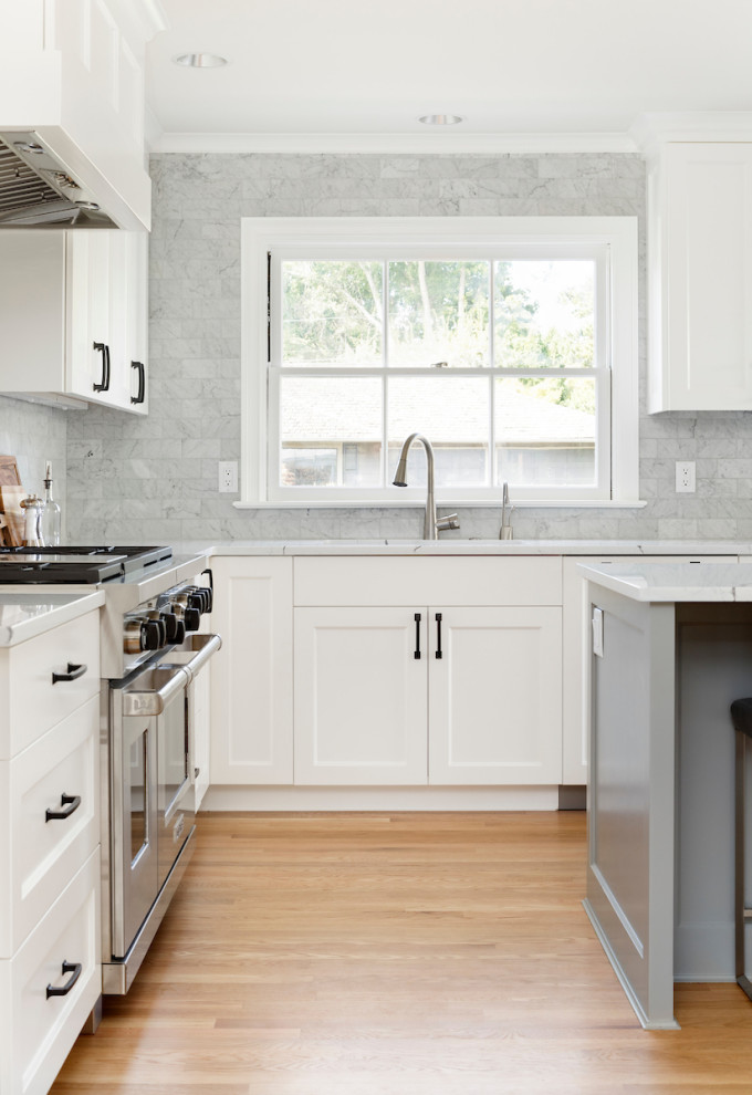 Bild på ett funkis vit vitt l-kök, med en undermonterad diskho, skåp i shakerstil, vita skåp, bänkskiva i kvarts, grått stänkskydd, stänkskydd i glaskakel, rostfria vitvaror, ljust trägolv, en köksö och brunt golv