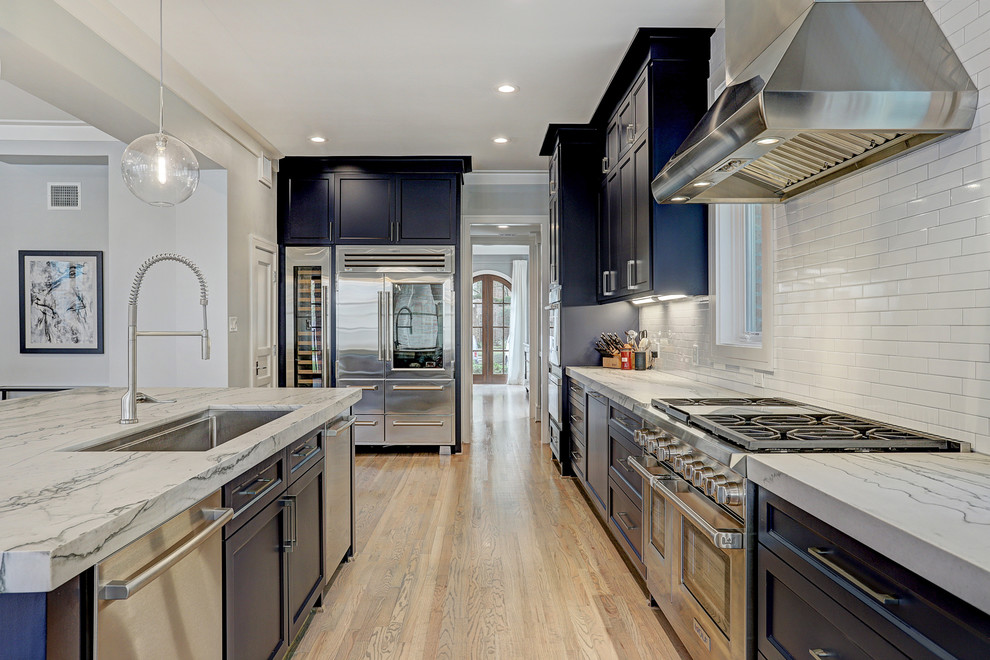 Photo of a large traditional l-shaped open plan kitchen in Houston with a built-in sink, recessed-panel cabinets, blue cabinets, quartz worktops, white splashback, metro tiled splashback, stainless steel appliances, light hardwood flooring, an island, brown floors and white worktops.